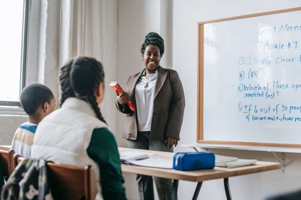 Happy black female teacher standing near whiteboard and explaining lesson to unrecognizable children sitting at wooden table with books and stationery